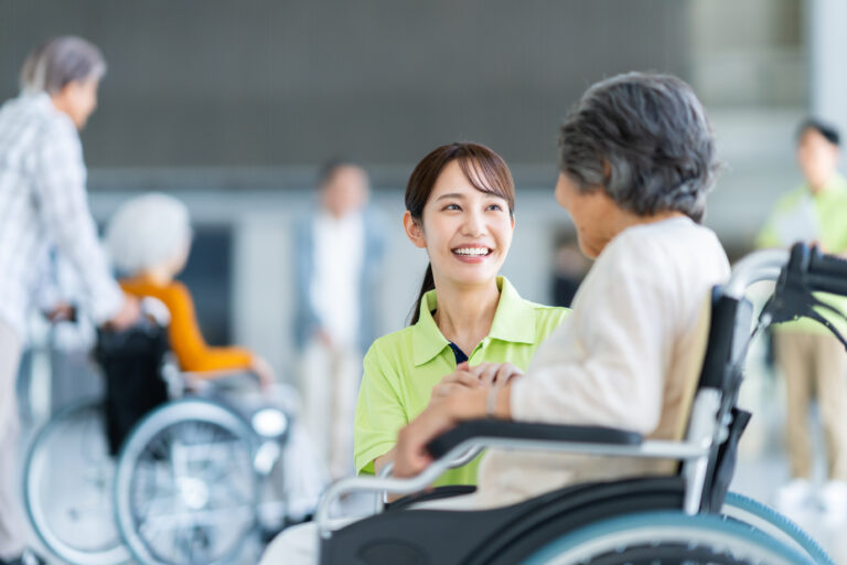 介護職員が入居者と交流する風景。複数の高齢者が車椅子で集まる共用スペース。